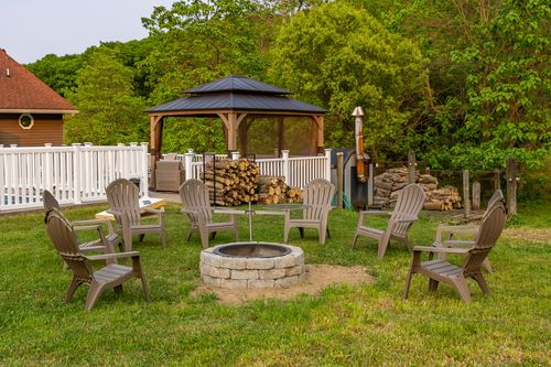 Circle of chairs around a stone fire pit near a gazebo and stacked firewood