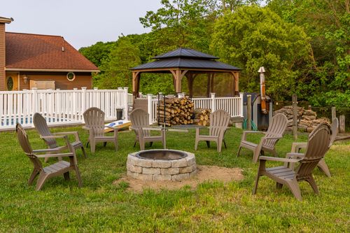 Circle of chairs around a stone fire pit with a cornhole board and firewood rack nearby
