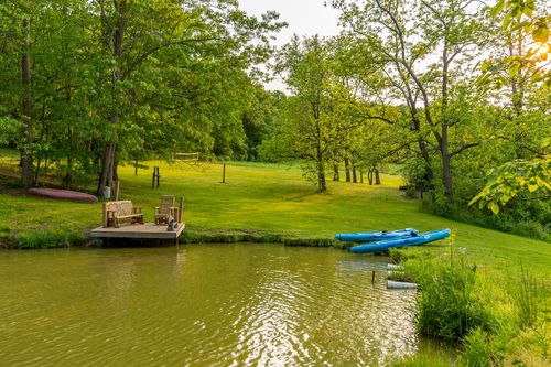 Wooden dock with bench seating beside a pond, kayaks and volleyball net on the lawn