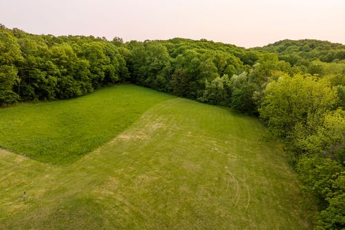 Aerial dusk view of a farmhouse with a pool and covered carport bordered by trees