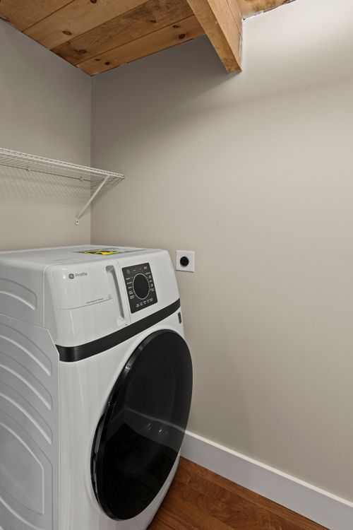 Laundry corner with a white front-load dryer beneath a wood ceiling beam and wire shelf