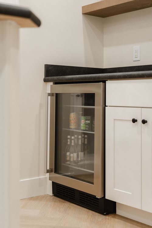 Built-in glass-front beverage refrigerator stocked with drinks beneath a floating wood shelf
