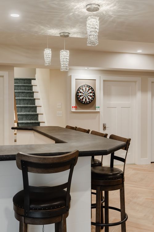 Basement bar with an L-shaped counter, dartboard cabinet and a carpeted staircase nearby