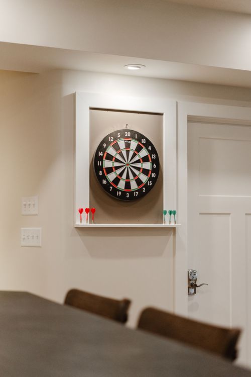 Wall-mounted dartboard in a white recessed cabinet with darts on the ledge