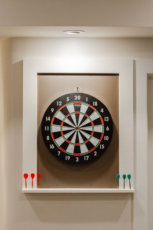 Recessed white cabinet framing a dartboard with red and green darts on the ledge