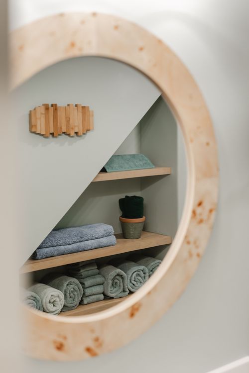 Round mirror reflecting under-stair shelving stacked with rolled towels