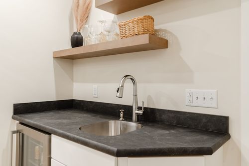 Close view of a wet bar's black stone counter, stainless sink and floating wood shelves