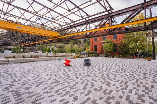 Open paver plaza with sculptural seating beneath a steel trestle and brick building