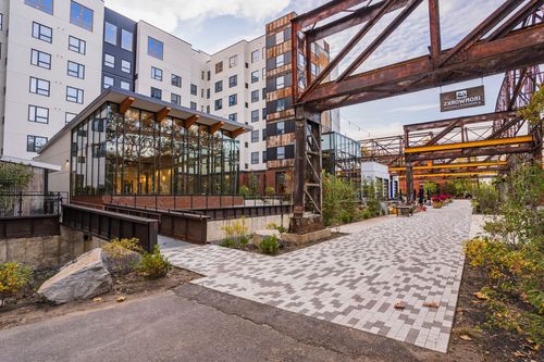 Paver walkway leading to a glass entrance with a hotel tower and steel trestle overhead