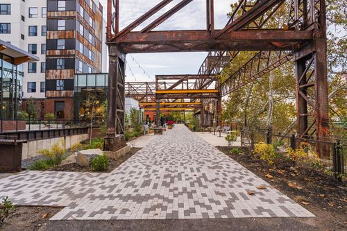 Paver walkway under a steel trestle structure with string lights and planting beds