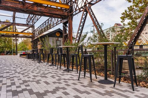 Paver plaza with black bar-height tables and stools beneath a steel trestle