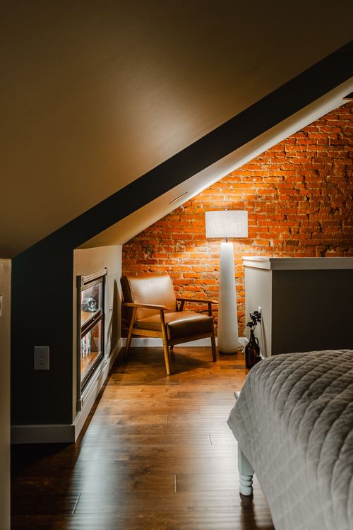 Bedroom at dusk with a beamed sloped ceiling and a warmly lit brick wall