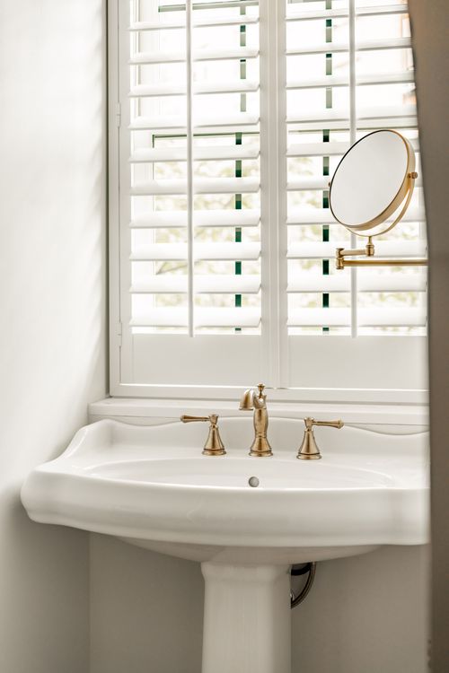 Pedestal sink beneath a plantation shutter window with brass faucet fixtures