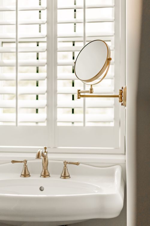 Close-up of a brass swing-arm makeup mirror beside a bathroom window
