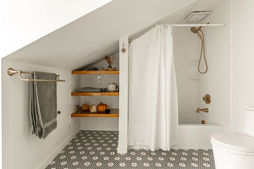 Attic bathroom with a white shower curtain, brass fixtures and wood shelving styled with pumpkins