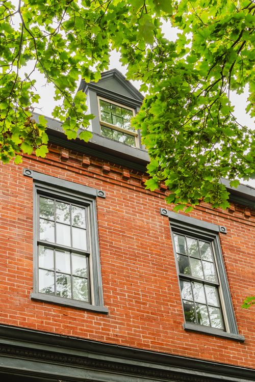 Upward view of a brick townhome facade with a dormer window framed by green tree canopy