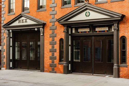 Twin arched entrances labeled REX and a wreath emblem set into a black-trimmed brick facade