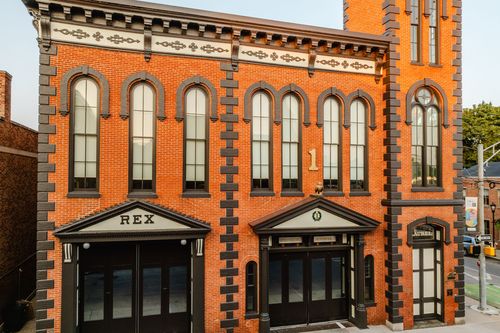 Street-level view of an arched-window brick facade with REX signage over black entry doors