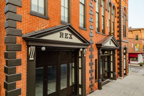 Close-up of a black-trimmed brick entrance with a REX pediment above glass storefront doors