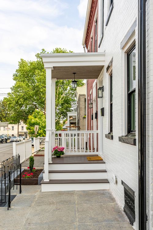 Covered front porch with white railings, wood steps and potted flowers along a city sidewalk