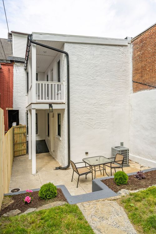 Rear white stucco addition with a second-floor balcony, patio table and garden beds below