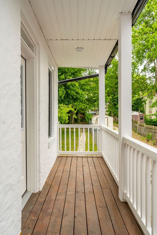 Covered wood-plank porch with white railings overlooking green trees and a backyard path
