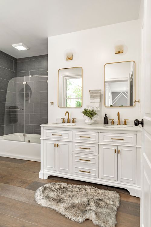 Bathroom with a white dual vanity, gold-framed mirrors and a gray tiled tub surround