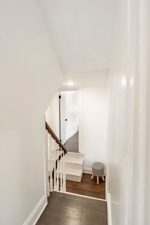 Narrow stairwell with white paneled walls leading down past a small round ottoman