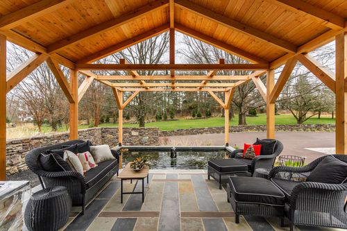 Vaulted timber-frame pergola ceiling over black wicker sofas beside a small pond