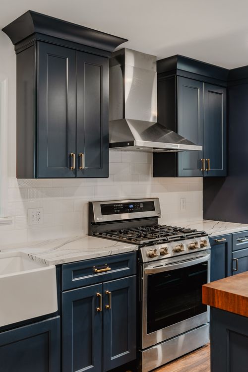 Navy cabinets with brass hardware beside a stainless range hood and subway tile