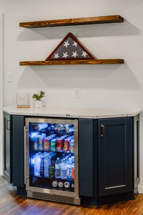 Stocked glass-front beverage fridge under floating shelves holding a folded flag case