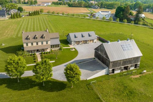 Aerial view of a farmhouse with wraparound porch beside a weathered barn with solar panels