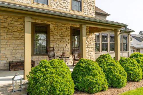 Covered stone porch with rocking chairs, an antique trunk and manicured shrubs