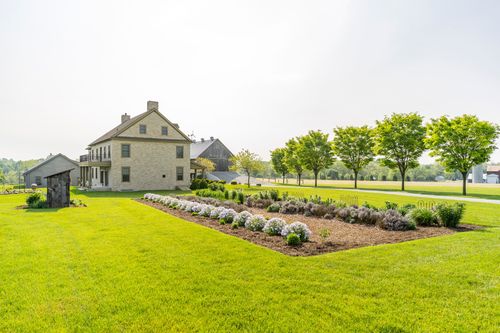 Stone farmhouse exterior beside a flowering garden bed and shade trees