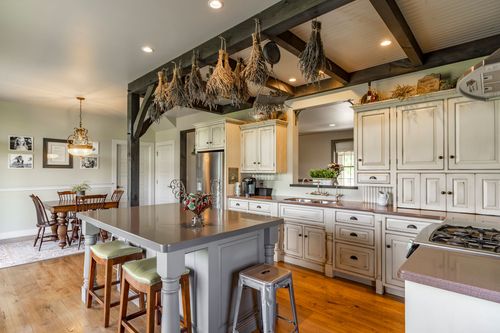 Farmhouse kitchen island with iron stools, hanging herbs and a dining table beyond