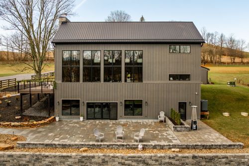 Rear exterior of a gray board-and-batten venue with tall black-framed windows and a stone-paved patio
