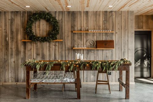 Rustic serving table strung with garland and lights beneath open glass shelving and a wreath