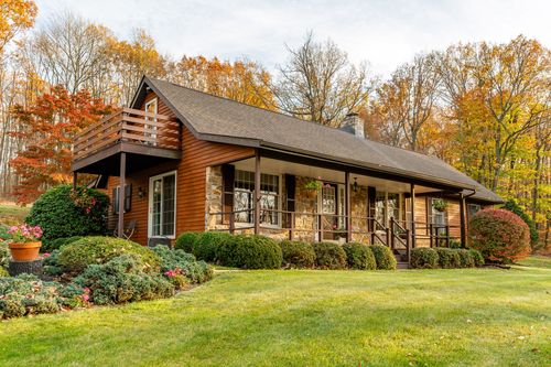 Log-style home with a wraparound porch, stone accents and a second-floor balcony amid fall trees