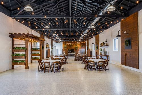 Banquet hall set with round tables, white linens, and wood chairs beneath a greenery-draped wood pergola