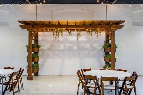 Wood pergola ceremony arbor with hanging greenery against a white brick wall