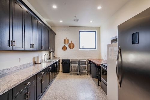 Catering kitchen with dark cabinetry, quartz counters, a stainless fridge, and an exposed brick wall