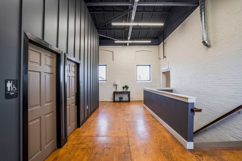 Hallway with restroom doors and a reception desk beneath arched brick windows and wood flooring