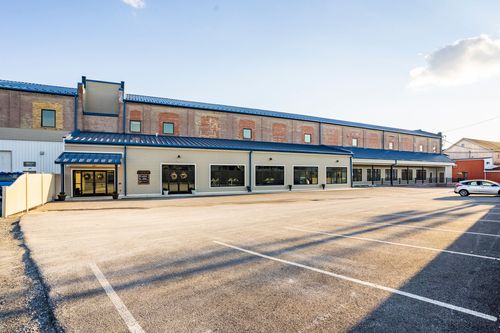 Ground-level view of a brick and tan mill building with a blue awning entrance and parking lot