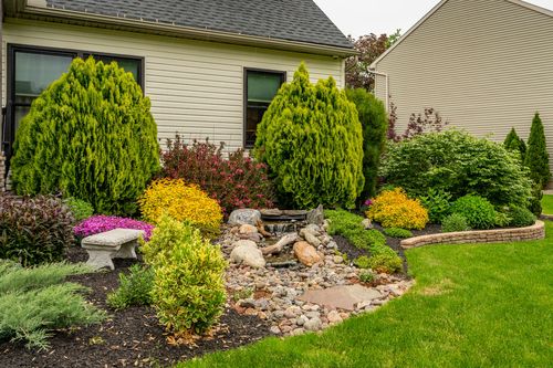 Landscaped side garden with manicured arborvitae, flowering shrubs, and a stone bench