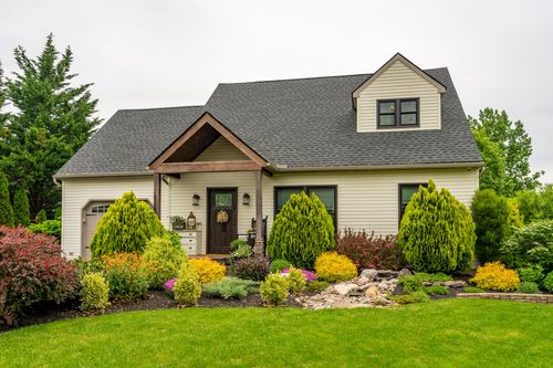 Cream cape-style home with gray shingle roof, wood gable entry, and lush landscaped foundation beds