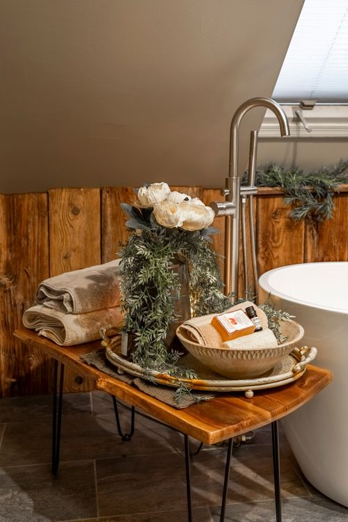 Close view of a floor-mount tub filler and a wood side table styled with towels and florals