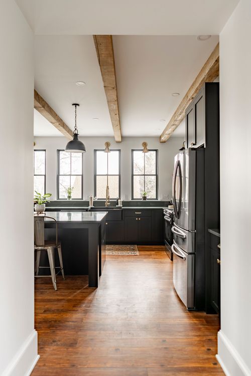 Hallway view into a black kitchen with wood beams, island and stainless refrigerator