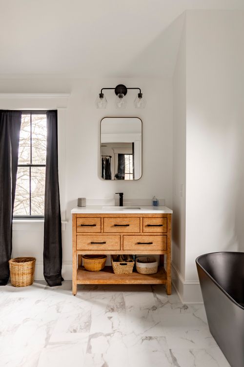 Wood bathroom vanity with a round mirror, black sconce and black soaking tub nearby