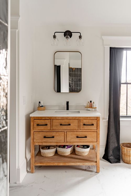Wood bathroom vanity with a brass-trimmed mirror below a black-curtained window