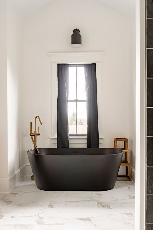 Black oval soaking tub with brass fixtures under an arched window with black curtain